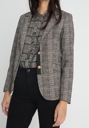 Vrouw die een grijs geruit blazer met gouden knopen draagt over een patroon knoopblouse en zwarte broek, staand tegen een effen achtergrond.