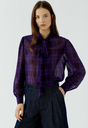Frau mit langen braunen Haaren, die eine transparente lila karierte Bluse mit großer Schleife und hoch taillierte dunkelblaue Hose trägt, steht mit einer Hand in der Tasche.