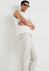 Witte mouwloze shirt met kleine graphic, lichtbeige broek en gele zonnebril, staande met gekruiste armen tegen een effen achtergrond.
