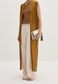 Długi, przejrzysty musztardowy kamizelka nałożona na brązowy podkoszulek w paski, w połączeniu z kremowymi spodniami typu wide-leg i jasnoszarym kopertówką z frędzlami.
