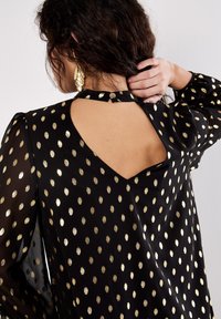 Schwarze Bluse mit goldenen Punkten, durchsichtige lange Ärmel, Schlüssel-Ausschnitt hinten mit Druckknopfverschluss, glatte Textur.