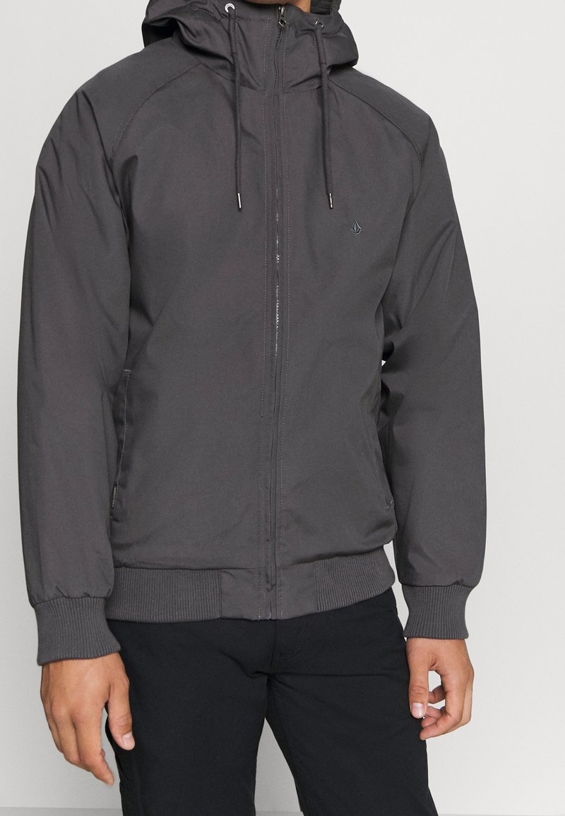 Hombre vistiendo una chaqueta con capucha de color gris oscuro, con cierre frontal y puños acanalados, combinado con pantalones negros frente a un fondo liso.