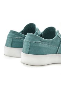 Turquoise canvas sneakers met een textuurafwerking, witte rubberen zolen, contrasterende stiksels en een afgerond achterkantdesign. Twee oogjes zichtbaar.
