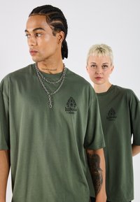 T-shirts oversize vert olive en tissu doux, avec un design de logo sur la poitrine ; chaînes en argent empilées sur un modèle, cheveux courts sur les deux.