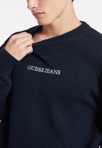 Sötétkék kötött pulóver bordás gallérral és mandzsettákkal. Az elején hímzett "GUESS JEANS" felirat található. Puha tapintású és laza fazonú.