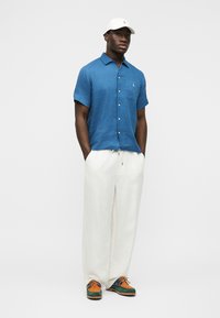 Vīrietis, kurš valkā zilu īsroku krekla kreklu ar pogām, baltas plašas bikses, daudzkrāsainas kurpes un balto sporta cepuri, stāv ar rokām kabatās.