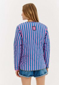 Kurtka pikowana w pionowe różowe i niebieskie paski, z okrągłym dekoltem i czarną metką na górnej części pleców. Spodnie dżinsowe widoczne poniżej.