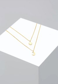Guldtonet lagdelt halskæde med en solstrålecharms og en halvmånecharms på fine kæder, sat mod en minimalistisk baggrund.