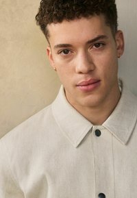 Jongere met kort krullend haar, die kleine oorringen draagt en een lichtbeige knopenjack aanheeft, voor een textuurmuur achtergrond.