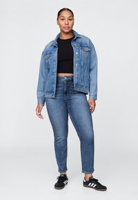 Šviesiai mėlyna džinsinė striukė su klasikiniu dizainu, su sagomis priekyje, krūtinės kišenėmis, derinama su tamsiai mėlynais aptemptais džinsais ir juodais sportbačiais.