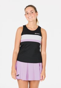 Giovane donna sorridente, che indossa un top sportivo senza maniche a righe bianche e nere e una gonna da tennis plissettata lilla, in piedi contro uno sfondo bianco.