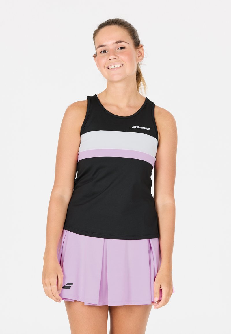 Giovane donna sorridente, che indossa un top sportivo senza maniche a righe bianche e nere e una gonna da tennis plissettata lilla, in piedi contro uno sfondo bianco.