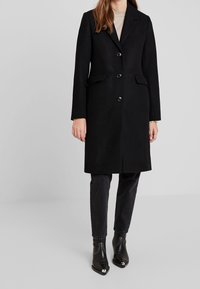 Schwarzer Wollmischmantel mit taillierter Passform, Revers mit Kerbkragen, drei vorderen Knöpfen und seitlichen Taschen. Getragen mit dunklen Jeans und schwarzen Stiefeletten.