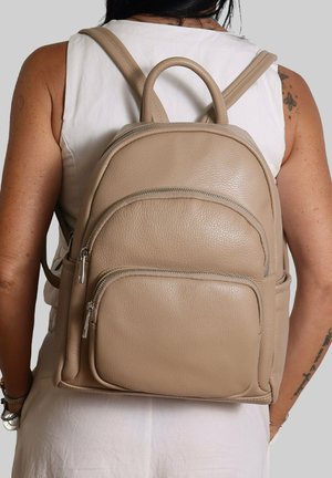 Persoon met een beige leren rugzak met twee met ritsen afgesloten vakken, een wit mouwloos topje en zichtbare tatoeages op de arm.