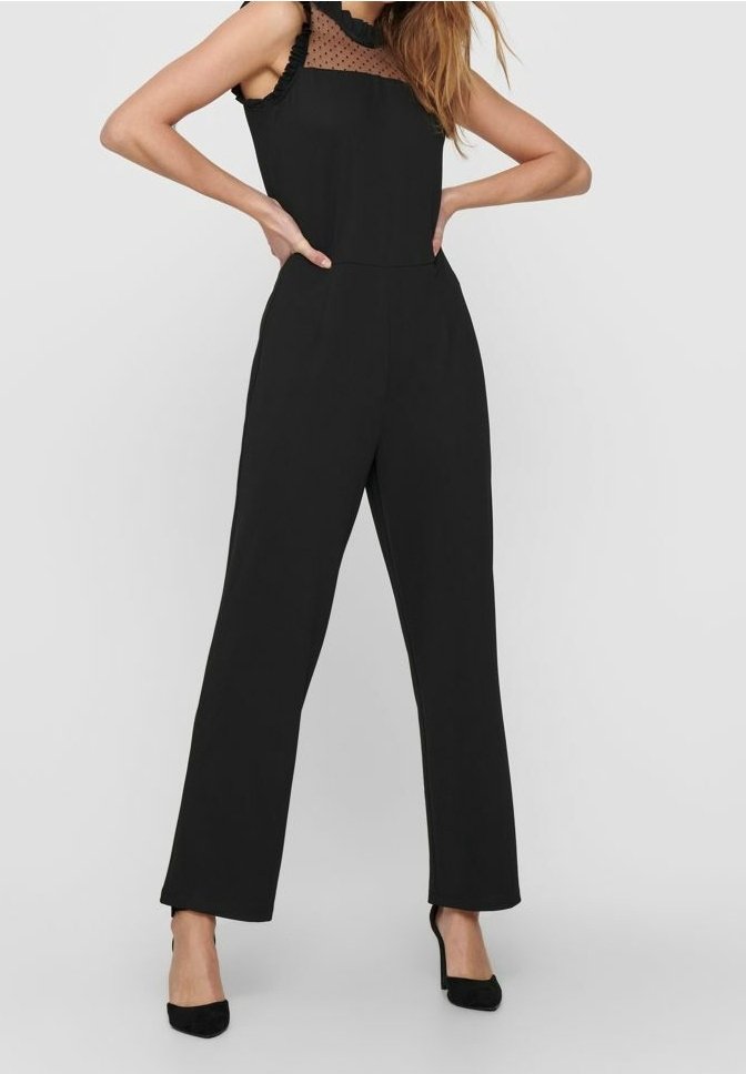 Combinaison noire avec un corsage ajusté, des épaules à volants et un empiècement transparent à pois. Pantalon à jambes larges, texture lisse, associé à des talons noirs.