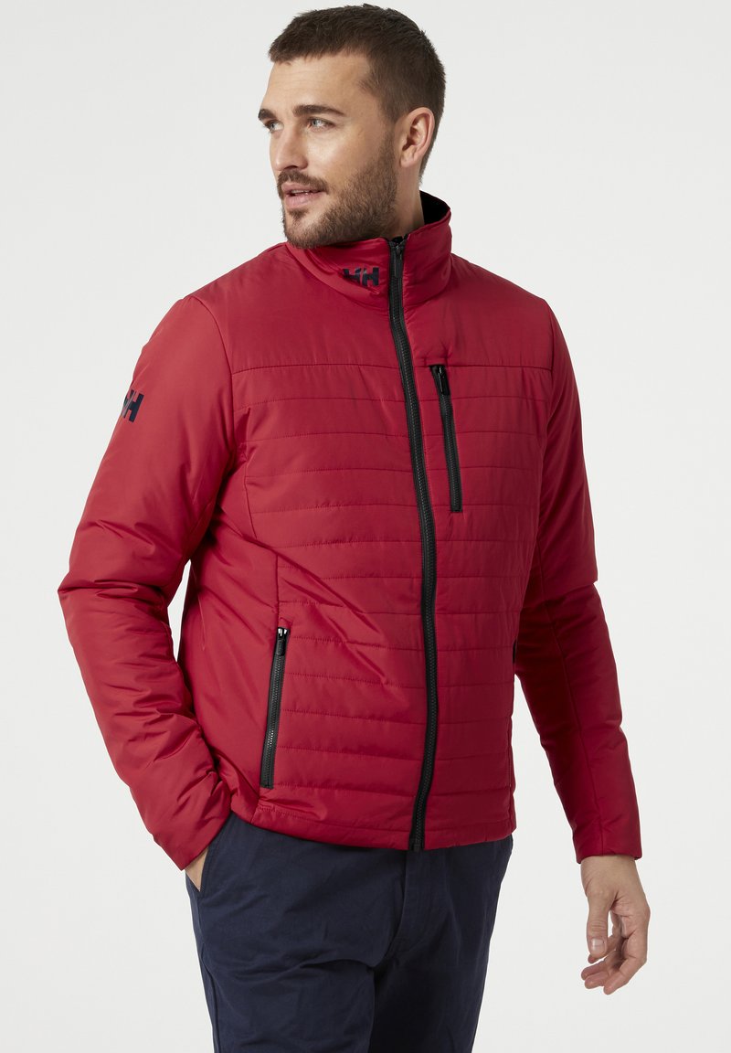 Muž ve červené prošívané bundě s černými zipovými kapsami a tmavě modrými kalhotami, hledící vlevo na jednobarevném pozadí.