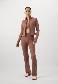 Brun idrettsett med en tettsittende glidelåsjakke og høyt midjebelte leggings, laget av et glatt, elastisk materiale. Sko med røde detaljer.