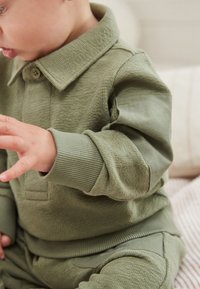 Olīvu zaļu mazuļa apģērbs ar strukturētu audumu, kas veidota ar apkaklīti, ribotām aprocēm un mīkstu iekšpusi.