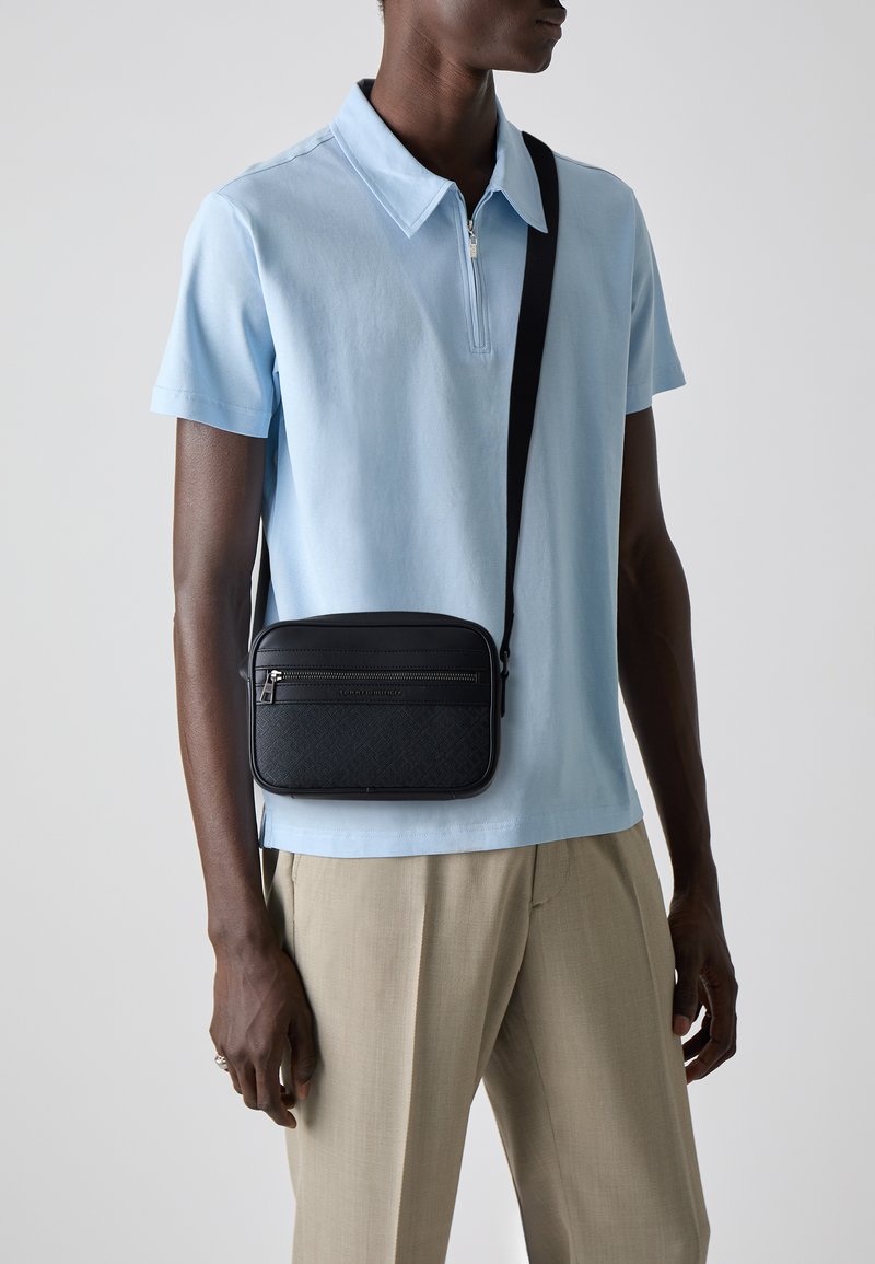 Sac bandoulière rectangulaire noir avec fermeture éclair à l'avant, surface texturée, bandoulière ajustable, associé à une chemise polo bleu clair et un pantalon beige.