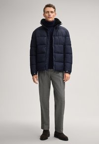 Giubbotto imbottito blu marina con sezioni trapuntate e collo foderato in pelliccia sintetica, abbinato a pantaloni grigi texture e stivali in suede marrone.