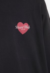 Czarna tkanina z małą grafiką czerwonego serca i białym napisem "Heart Thief since 1991" umieszczonym na środku.