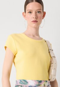 T-shirt jaune à côtes avec un col rond et des manches courtes. Présente un détail brodé subtil. Assorti à une jupe multicolore et à une sangle beige.