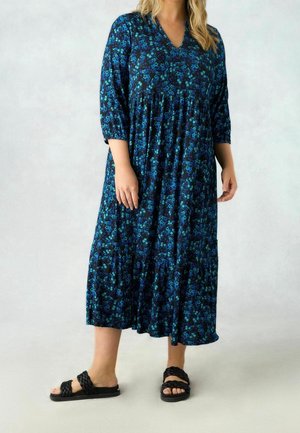 Vrouw draagt een blauw-zwart gebloemde midi-jurk met driekwart mouwen en zwarte gevlochten slides, staande op een lichte achtergrond.