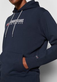 Personne portant un sweat à capuche Tommy Jeans bleu marine avec cordons blancs, poche avant et texte du logo « Tommy Jeans New York City » sur la poitrine.