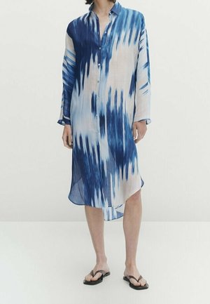 Femme portant une robe chemise mi-longue à manches longues avec des motifs abstraits bleus et blancs, ainsi que des sandales tongs noires.