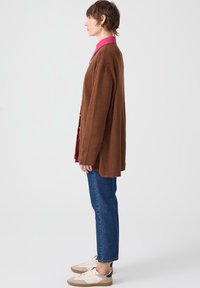 Brown corduroy cardigan with oversized fit, layered over a pink shirt. Blue jeans and white sneakers with beige accents complete the look.