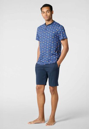 Uomo in piedi a piedi nudi che indossa una camicia blu con maniche corte e bottoni, decorata con stampe di uccelli, e pantaloncini blu scuro, su sfondo semplice.