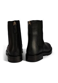 Ein Paar schwarze Lederstiefeletten mit hinteren Reißverschlüssen, elastischen Seitenpaneelen und niedrigen, gestapelten Absätzen auf einem weißen Hintergrund.