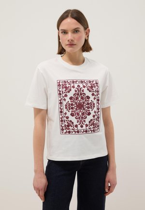 Mujer con cabello corto castaño que lleva una camiseta blanca con un diseño cuadrado intrincado de flores y enredaderas en tonos rojo oscuro, combinada con jeans oscuros.