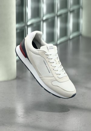 Hvid Tommy Hilfiger-sneaker med navyblå og røde detaljer, hvide snørebånd og mærket tekst på siden og tungen, ophængt over en grå gulv.