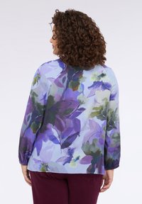 Blusa floreale con maniche lunghe, caratterizzata da uno sfondo blu e motivi di foglie in acquerello viola-verdi; tessuto morbido e leggero con polsini elasticizzati.