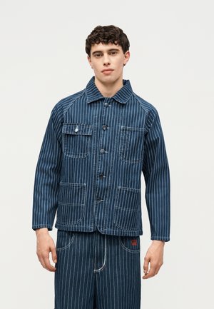 Junger Mann trägt eine blaue Nadelstreifenjacke mit vier Vordertaschen und passenden Nadelstreifenhosen vor einem schlichten Hintergrund.