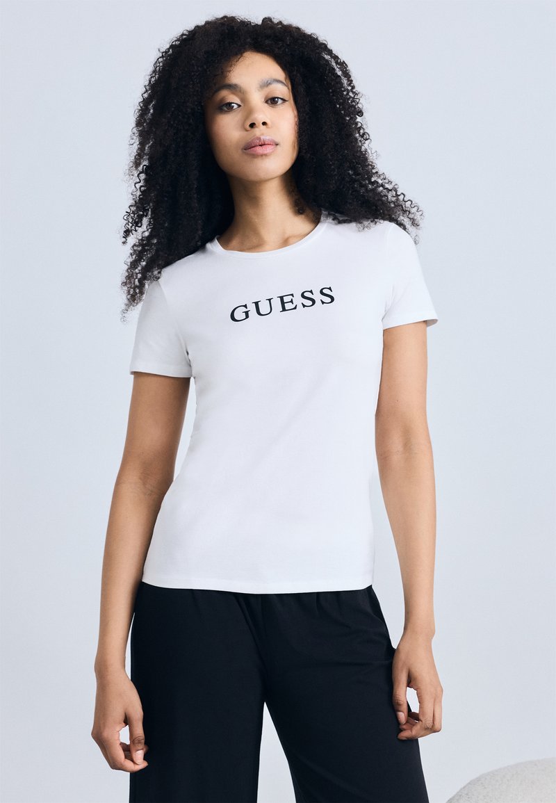 White cotton t-shirt with short sleeves, featuring the word "GUESS" in bold black letters across the chest. Smooth texture and classic fit.