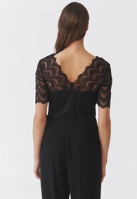 Sort top med korte blonder ærmer, der har en bølget kant og et V-ryg design. Glat stofkrop med en tætsiddende silhuet.