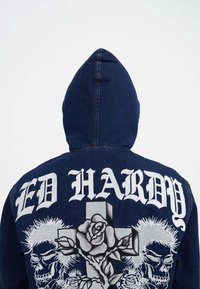 Jeansjacke mit Kapuze; dunkelblaue Farbe. Verfügt über großen weißen Grafiktext "ED HARDY" und kunstvoll gestickte Skull- und Rosenmuster auf dem Rücken.