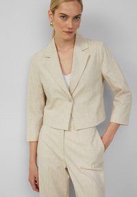 Blazer court beige avec des manches trois-quarts, revers à cran, fermeture à un bouton et un motif texturé subtil.