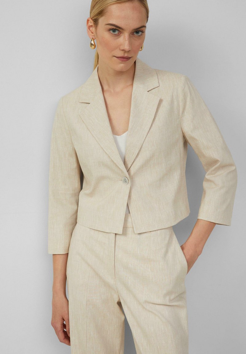 Blazer court beige avec des manches trois-quarts, revers à cran, fermeture à un bouton et un motif texturé subtil.