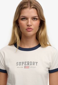 Roomwit T-shirt met korte mouwen en een marineblauwe kraag, voorzien van een opdruk 'SUPERDRY' en een ontwerp van de Amerikaanse vlag. Zachte katoenen stof, klassieke pasvorm.