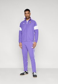 Μωβ φόρμα με λευκές λεπτομέρειες, που περιλαμβάνει ζακέτα με φερμουάρ και εφαρμοστά παντελόνια. Περιλαμβάνει το λογότυπο Adidas και τρεις ρίγες στα παντελόνια.