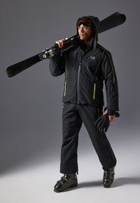 Chaqueta y pantalones de esquí negros con acentos amarillos, sosteniendo esquís. Incluye gafas, guantes y botas de esquí. Material impermeable y aislante.