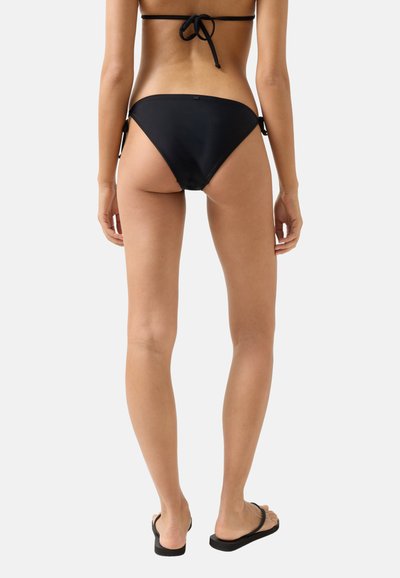 Frau, die mit dem Rücken zur Kamera steht, trägt schwarze Bikinihosen und Flip-Flops vor einem schlichten hellen Hintergrund.