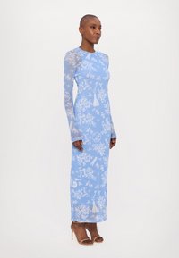 Robe bleu à manches longues avec un motif floral, une texture transparente et une silhouette droite, associée à des talons beiges à lanières.