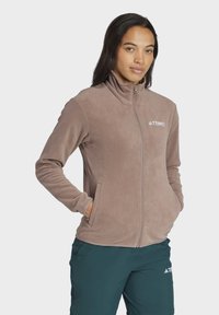 Fleecejacke in Hellbraun, mit hohem Kragen, durchgehendem Reißverschluss, Seitentaschen und einem weißen Logo auf der Brust zur Markenkennzeichnung.