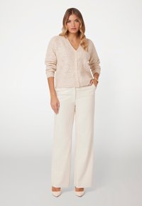 Cardigan lavorato a maglia di un beige chiaro con scollo a V, caratterizzato da motivi testurizzati e chiusura con bottoni. Abbinato a pantaloni a gamba larga color crema e tacchi beige.