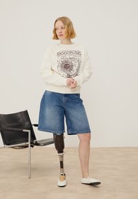 Jeune femme aux cheveux courts portant un sweat à motifs à manches longues et un short en jean, debout avec une jambe prothétique près d'une chaise noire.