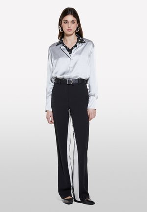 Camicia grigia in seta con colletto nero, abbinata a pantaloni neri con strisce laterali bianche e una cintura nera. Scarpe basse completano il look.